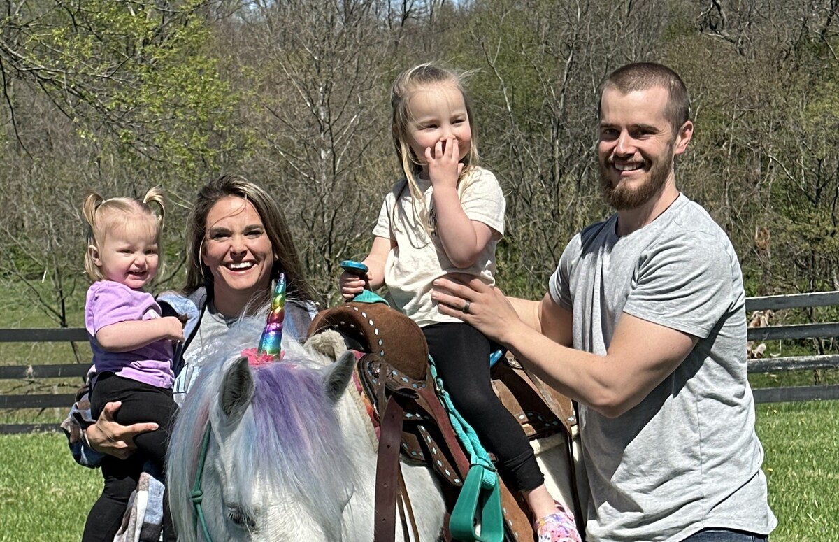 A smiling family consisting of a man, a woman, and two young girls, one of whom is on a horse